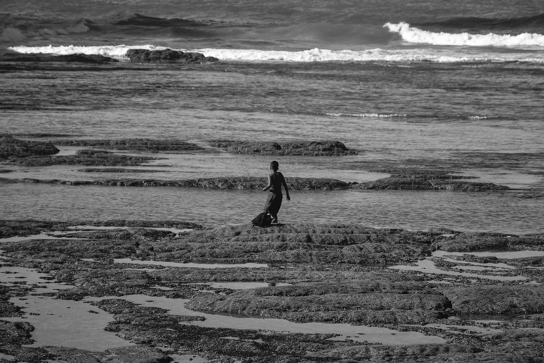 A black and white image of a person walking along the rocky seashore in Casablanca, Morocco.