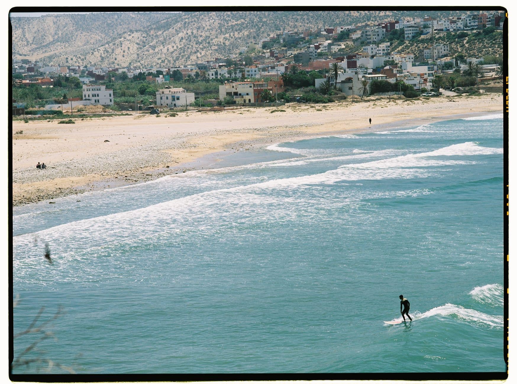 Aerial view of a surfer catching waves along the scenic Moroccan beach with mountains in the background.