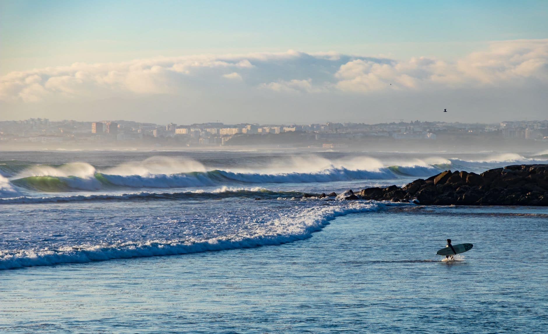 A surfer approaches ocean waves at Costa da Caparica beach with a clear sky backdrop.