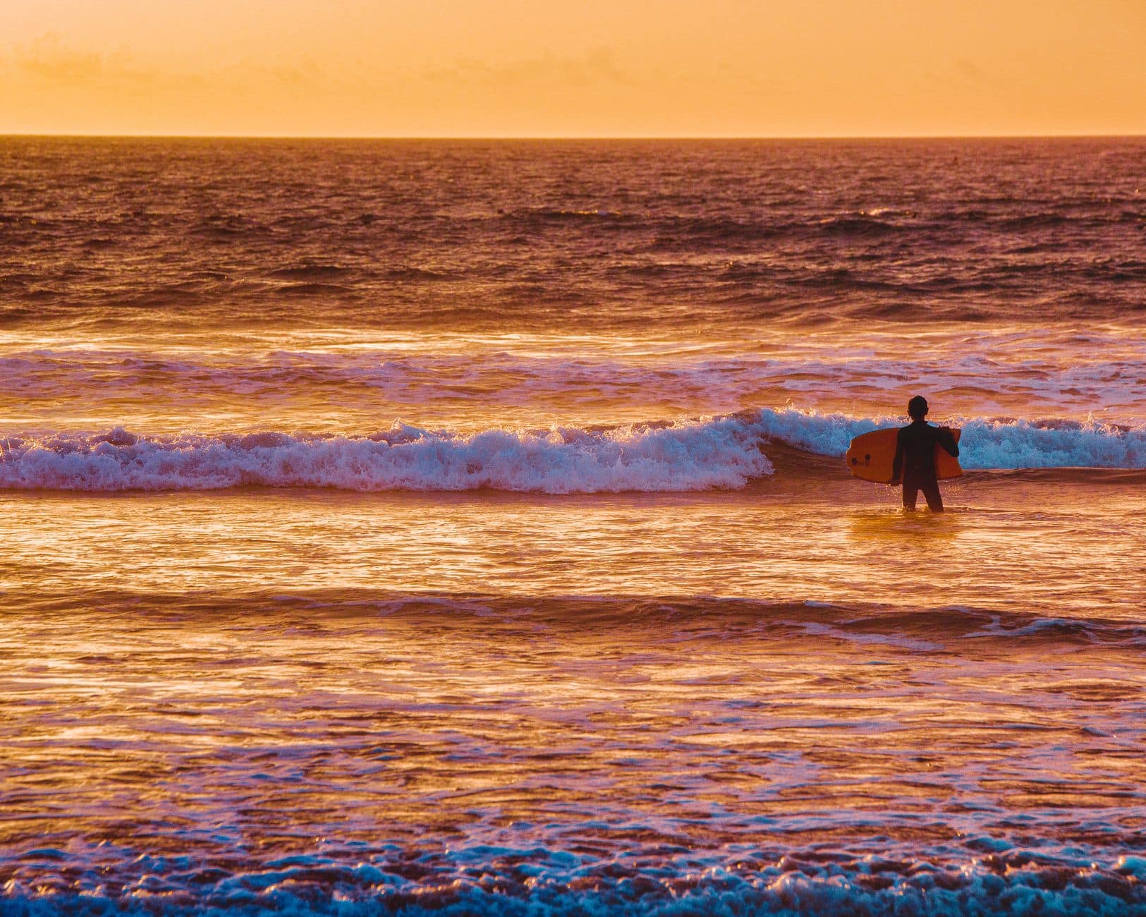 A silhouette of a surfer entering the ocean at sunset on Costa da Caparica beach, Portugal.